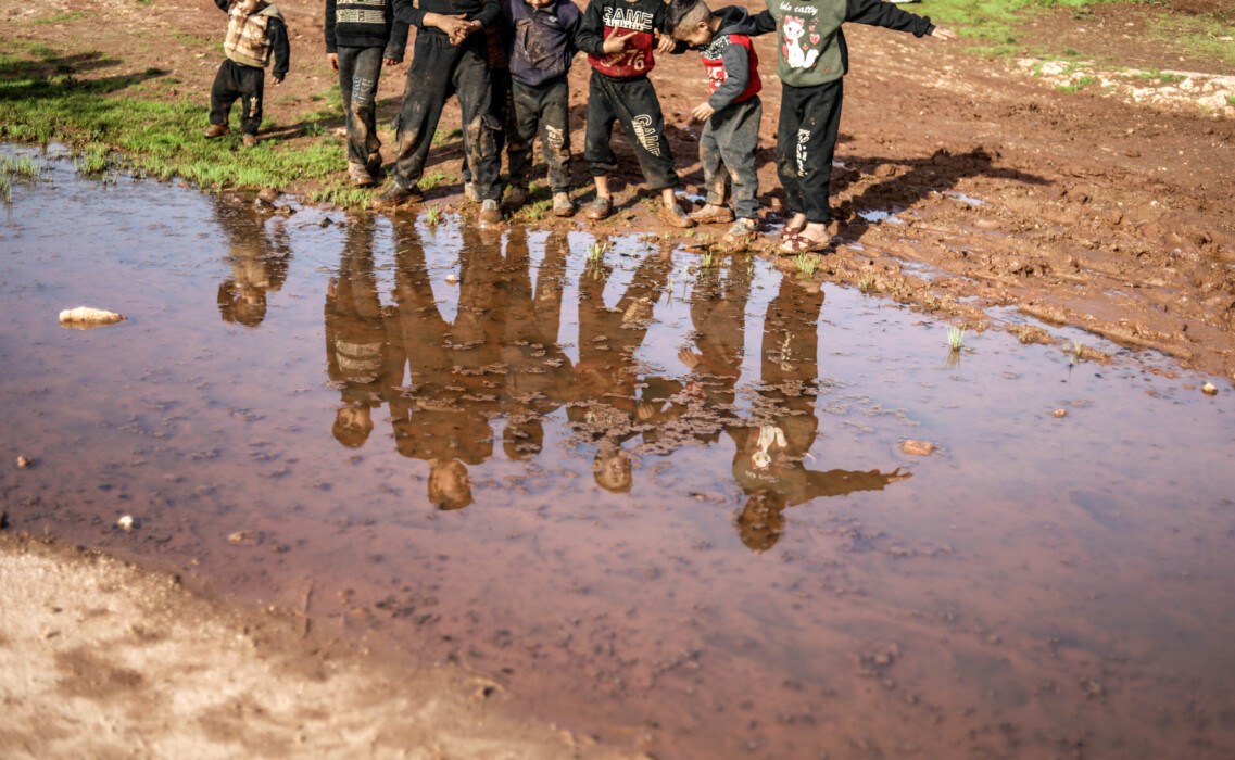 IDLIB, SYRIA - JANUARY 20: Syrian children, who have to struggle with hard life conditions and cold winter as refugees in Bettin Camp, walk in mud by a puddle formed after the rainfall in Idlib, Syria on January 20, 2024. Due to the lack of infrastructure and sewerage network in the camp, the muddy stream formed as a result of the rainfall negatively affected the lives of the refugees by forcing them to pass through the puddles and muddy roads. (Photo by Izettin Kasim/Anadolu via Getty Images)